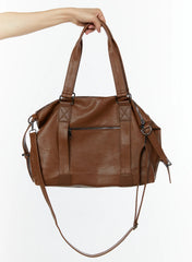 LEWKIN Unisex Leather Utility Bag IU515 in Brown, hand-held product shot showing front zip pocket, X-stitch hardware tabs, dual top handles, and removable crossbody strap.