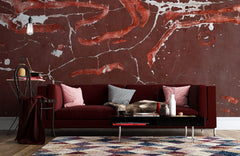 Rustic red abstract cracked earth mural wallpaper behind maroon sofa with geometric rug in living room.