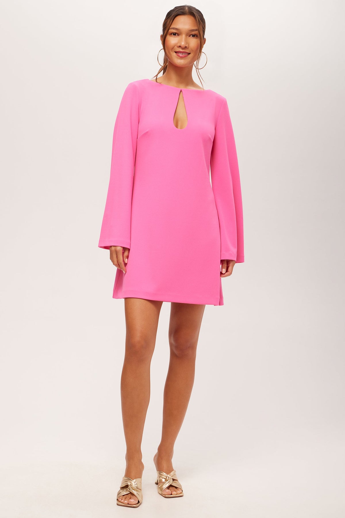 A woman stands against a light background wearing the Singles Long-Sleeve Keyhole Dress in Love All Pink, featuring a front keyhole detail. Her hair is tied back, and she accessorizes with large hoop earrings and gold open-toed sandals.
