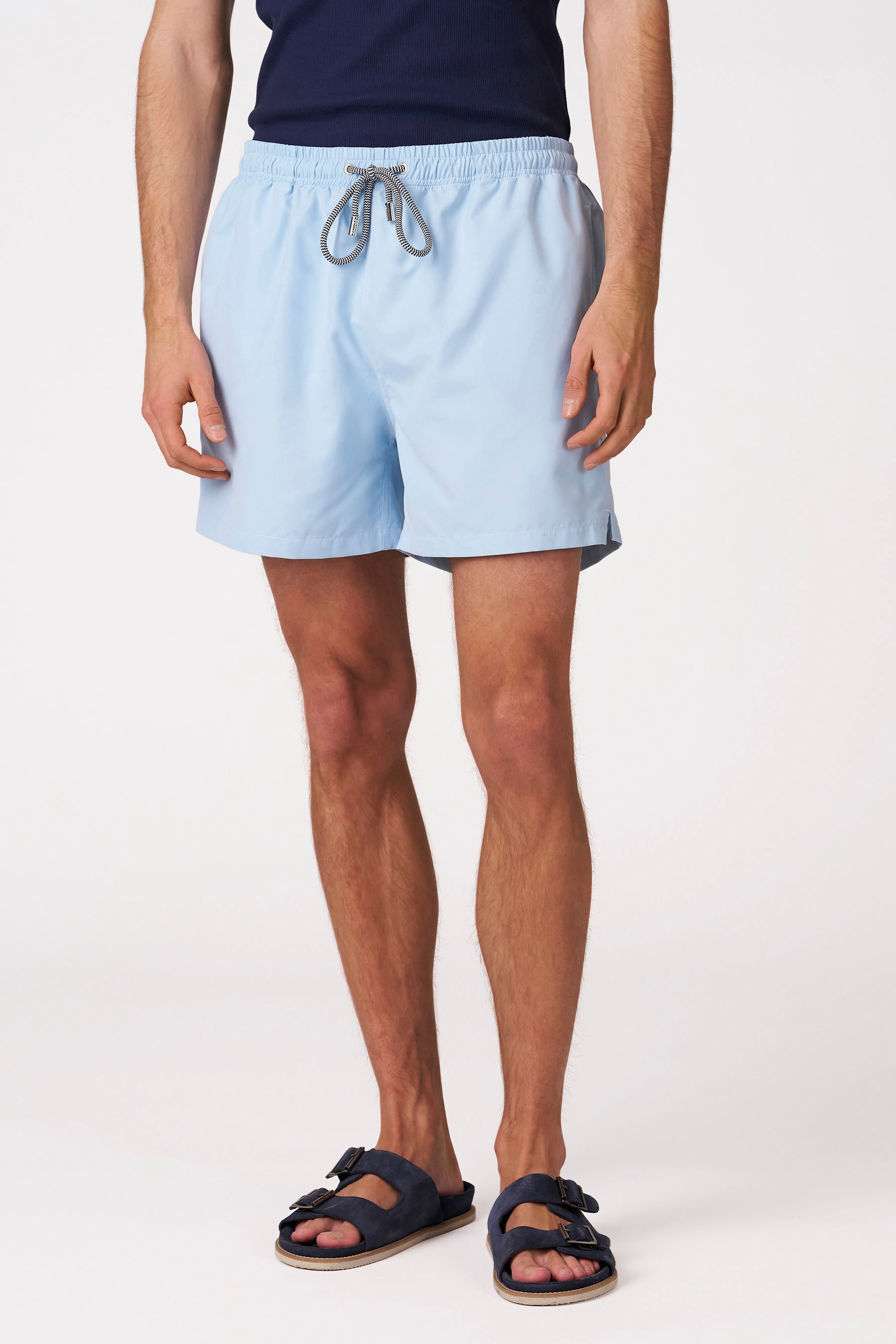 Blauwe zwemshort van Bruce & Butler met elastische tailleband, zwart-wit trekkoord en splitten aan de zijkant, gedragen door een man met donkerblauwe sandalen.