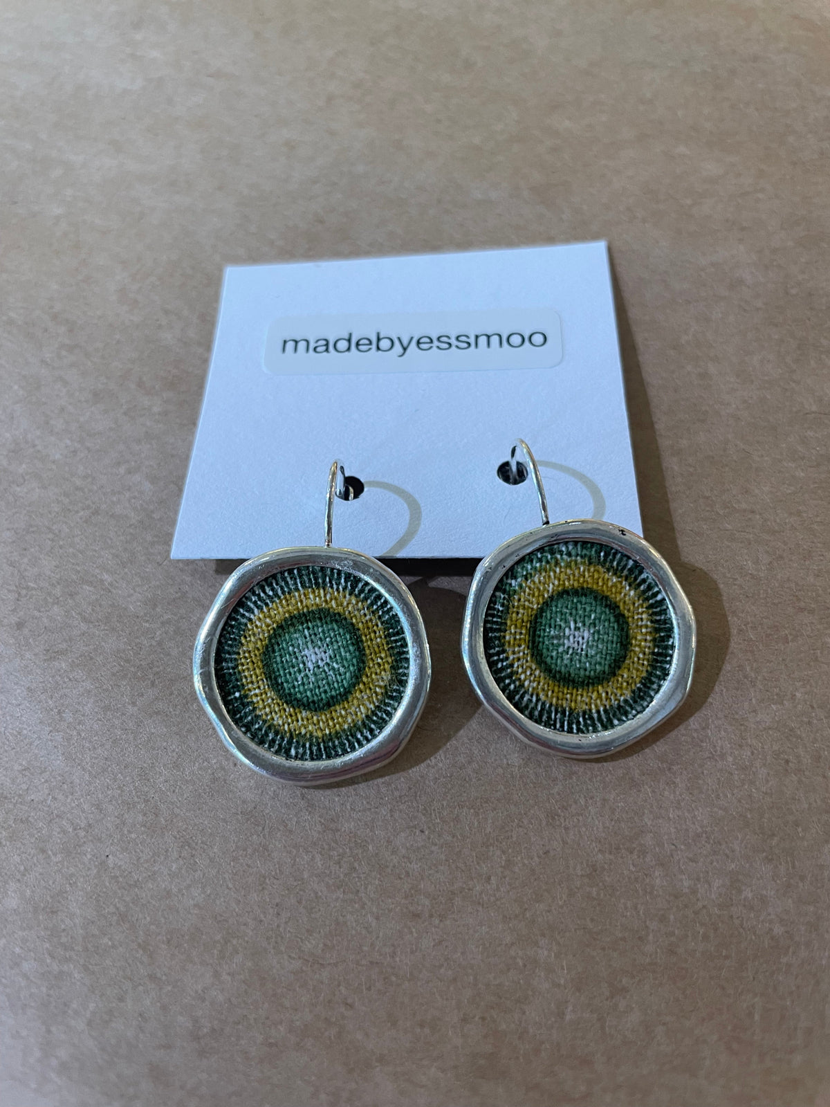 A pair of Essmoo earrings by BUTIK KUBIK, round and handmade with green, yellow, and black circular patterns, are displayed on a brown background with a madebyessmoo card.