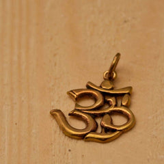 Ein Om-Anhänger aus Bronze liegt auf einer Holzoberfläche. Das filigrane Design betont die traditionellen stilisierten Kurven und Schlaufen des Om-Symbols, das in indischen Religionen für Klang und Spiritualität steht.