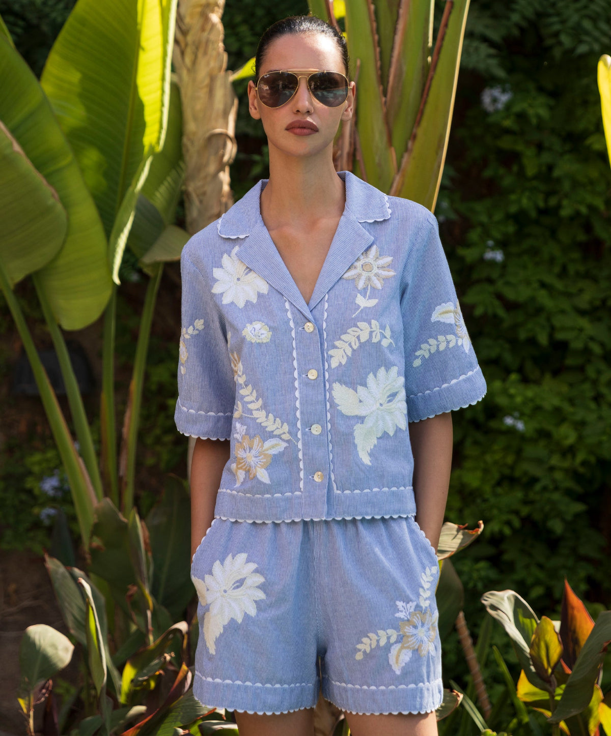 Woman wearing an embroidered blue striped shirt with matching shorts and sunglasses in a garden setting