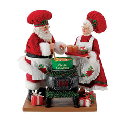 Santa and Mrs. Claus figurines in red chef outfits cooking together at a garland-wrapped stove with a green "Merry Christmas" pot