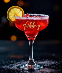 A personalized margarita glass with salt and lemon on the rim of the glass and a girls name in gold letters. Filled with red margarita and standing on a black table with salt around it.